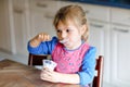 Little toddler girl eating yogurt for breakfast. Cute healthy baby sitting in the kitchen or at nursery and having meal.