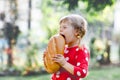 Little toddler girl holding big loaf of bread. Funny happy child biting and eating healthy bread, outdoors. Hungry kid. Royalty Free Stock Photo