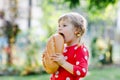 Little toddler girl holding big loaf of bread. Funny happy child biting and eating healthy bread, outdoors. Hungry kid.