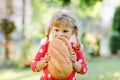 Little toddler girl holding big loaf of bread. Funny happy child biting and eating healthy bread, outdoors. Hungry kid. Royalty Free Stock Photo