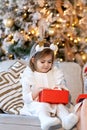 A little toddler girl sits on a sofa near a decorated Christmas tree and opens presents. Christmas Holidays Royalty Free Stock Photo