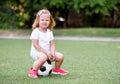 Little toddler girl in sports uniform and pink sneakers sitting on a soccer ball in a green football field outdoors with a funny Royalty Free Stock Photo