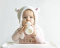 Little toddler girl in a warm fluffy hat drinks milk from a bottle while sitting. Half-length portrait. White gray