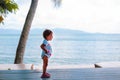 Little toddler girl in a white dress looks at the sea while standing on the wooden floor outside. back view