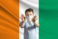 Little white boy in a protective mask on the background of the flag of Cote d`Ivoire Makes a stop sign with his hands, stay at