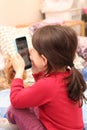 A little white girl with a ponytail takes a picture of her sister on a cell phone against the background of a regular apartment