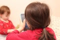 A little white girl with a ponytail takes a picture of her sister on a cell phone against the background of a regular apartment