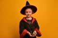 Cute little wizard girl holding toy spider and looking at camera on yellow background. A kid wearing Halloween costume.