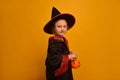 Little wizard kid girl with pumpkin candy basket looking at camera. Child in wizard robe and hat on a yellow background.