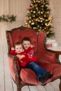 Litttle brother hugs his little sister in red retro chair near Christmas Tree. Enjoying a love hug, people`s holidays.