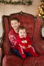 Litttle brother hugs his little sister in red retro chair near Christmas Tree. Enjoying a love hug, people`s holidays.