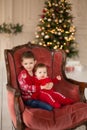 Litttle brother hugs his little sister in red retro chair near Christmas Tree. Enjoying a love hug, people`s holidays.