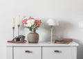 Living room interior - candles, fresh flowers in a ceramic vase, a table lamp, a notebook and a camera on a white chest of drawers