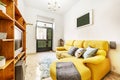 Living room of a rental house with a yellow sofa with a chaise longue, a wooden shelf and access to the street through a black