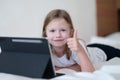 Llittle girl is lying on bed with tablet and holding thumbs up