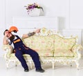 Loader sit on sofa, having rest. Man with beard, worker in overalls and helmet sits on couch tired, white background