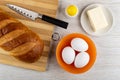 Loaf of bread, knife on cutting board, salt shaker, butter in saucer, chicken eggs in bowl on table. Top view Royalty Free Stock Photo