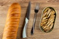 Loaf of bread, knife, fork, opened jar with canned sprats on wooden table. Top view