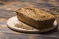 A loaf of freshly baked buckwheat bread lies on a round kitchen board on a brown wooden table. A handful of green