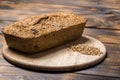 A loaf of freshly baked buckwheat bread lies on a round kitchen board on a brown wooden table. A handful of green