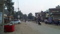 Local area street of a city area of country lndia where small buildings car people bike shop & trees are there