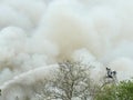 Firefighters up in bucket spraying a stream of water on an unseen fire, the background filled with white smoke