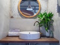 Loft style bathroom interior. Round mirror on concrete wall with brass faucet, white sink basin, green leaves in ceramic vase and