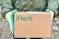 Logotype iHerb on cardboard box in courier hands. iHerb is an herbal products company