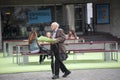 An elderly gray-haired man in a suit with a bouquet of flowers walks past a street cafe