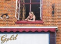 Woman leaning out upstairs window looking along street holding mobile phone