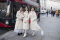 Three Chinese girls in white coats and dresses posing against the backdrop of a passing bus