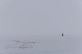 A lone fisherman sits on the ice in a thick fog
