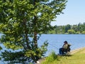 Lone fisherman sitting folding chair in shade of tree by lake