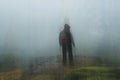 A lone ghostly, hooded hiker looking down from a hill, on a rainy, foggy day. With a dark, grunge, vintage edit