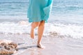 A lone girl in a blue dress walks along the waterline of a sandy beach. Women`s feet along the beach. The concept of travel,