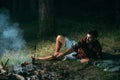 Lone hiker camping in forest. Handsome hipster cooking sausages over fire. Bearded barefoot man lying on grass, unity
