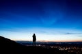 A lone hooded figure on a hill silhouetted just before sunrise looking out on city lights