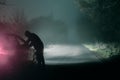 A lone hooded figure looking into a car. Silhouetted on a spooky, country foggy winters road at night