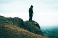 A lone sinister, hooded figure standing on a rocky outcrop looking out from top of a hill