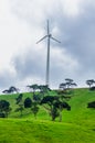 A lone windmill standing atop a distant mountain range with green grass pastures and trees in the foreground