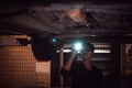 Lonely Asian male shining flashlight to examine car under chassis of automotive vehicle at old abandoned garage in night scene.