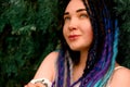 A lonely beautiful girl with multi-colored dreadlocks in a white dress is standing on a red stairs near, green cypress, thuja tree