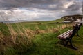 Lonely bench in a cold and windy coastal city, pointing to the sea with dramatic cloudy sky while grass is blown by the wind.