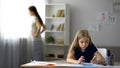 Lonely child drawing picture, strict mother standing by window, tense atmosphere