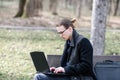 A lonely European student with glasses and a coat works on a laptop while sitting on a park bench
