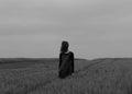 Lonely female standing in large windy grass field with her face covered with hair shot in greyscale Royalty Free Stock Photo