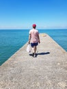 A lonely fisherman holding his fishing rod and a plastic bag and walking along a lonely pier, blue sea and sky in the