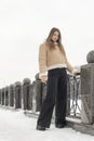 Lonely girl in beige sheepskin coat and black pants stands in snow-covered city park. Casual winter look for city Royalty Free Stock Photo