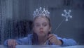 Lonely little princess in crown with magic stick sitting behind rainy window