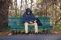 Lonely man in blue jacket, with hood dressed sits in autumn in the Park on green bench and reads book. there is black backpack and